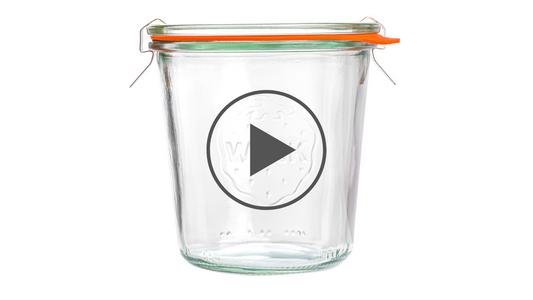 Стеклянная банка Weck с оранжевой уплотнительной резинкой и металлическими зажимами, с вытисненным логотипом на фронтальной стороне, охваченная кругом с символом воспроизведения видео, на белом фоне.