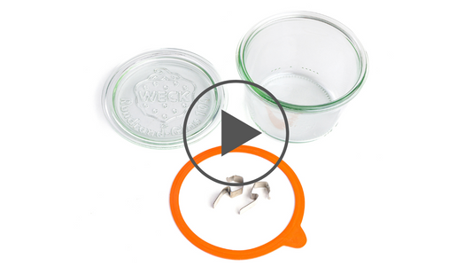 Открытая стеклянная банка Weck вид сверху с оранжевым силиконовым уплотнителем и металлическими зажимами, размещенными внутри прозрачного круга с символом воспроизведения видео, на белом фоне.