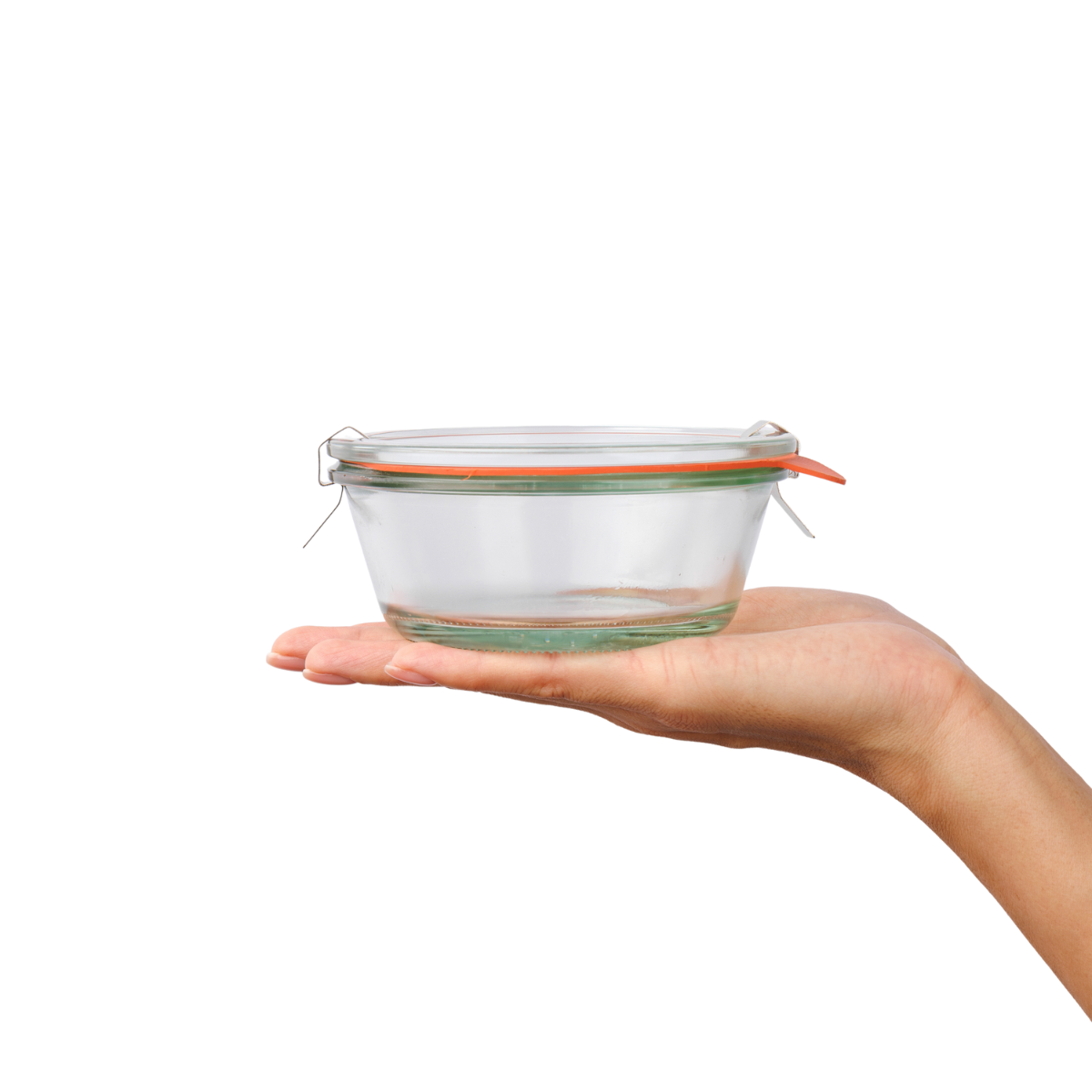 Ладонь держит прозрачную стеклянную банку WECK с закругленными краями и гладкими стенками, оранжевой уплотнительной резинкой и металлическими зажимами, с четкой гравировкой логотипа на боку.