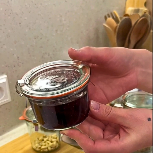 Закрытая стеклянная банка Weck 976 с джемом, которую держат на фоне деревянной разделочной доски и другого стеклянного банки.