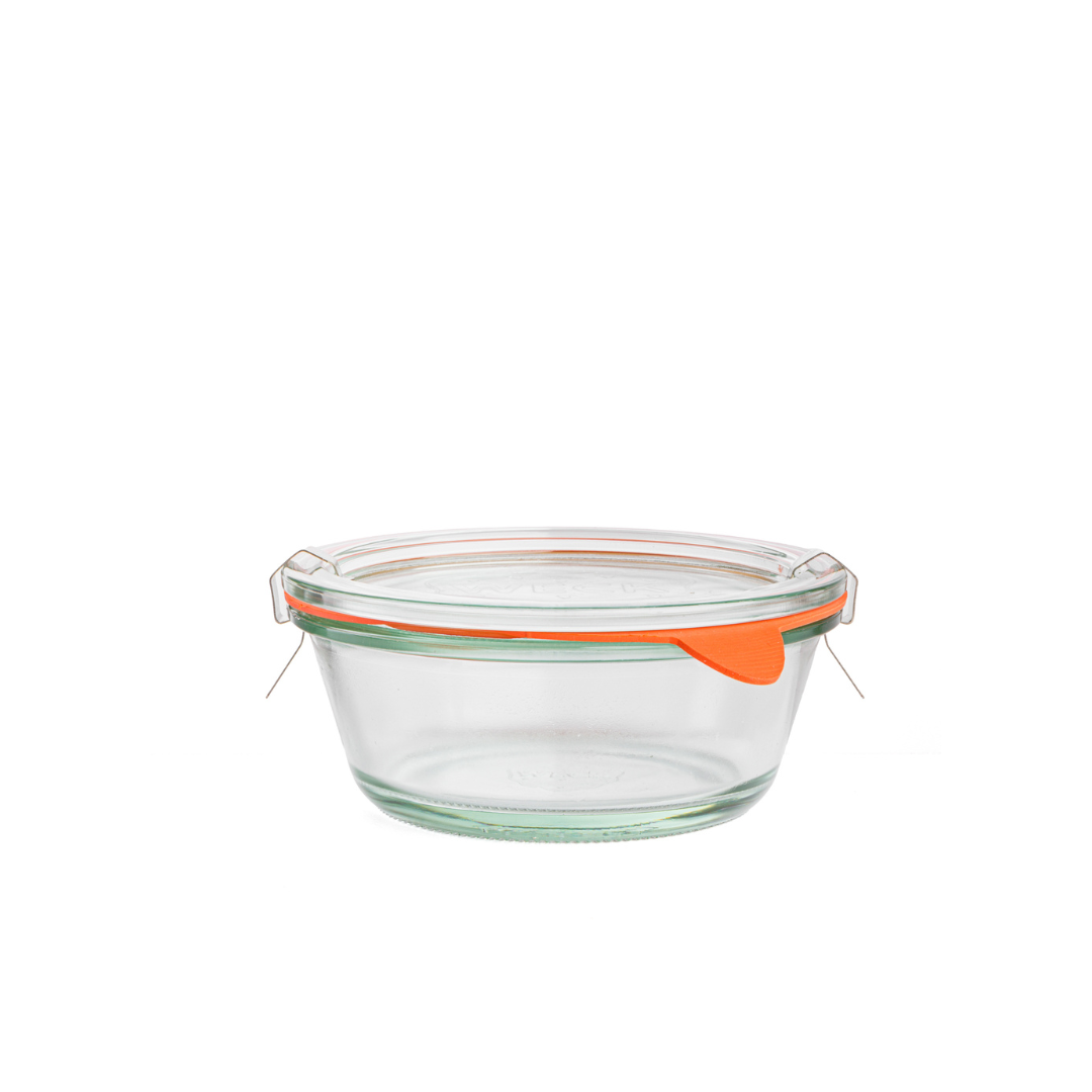 Круглая стеклянная банка Weck с тонкими стенками, прозрачной крышкой с гравировкой логотипа, оранжевым силиконовым уплотнителем и металлическими зажимами, на белом фоне.