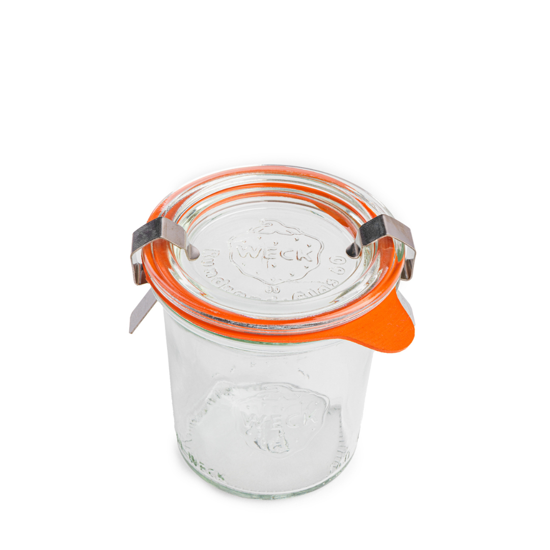 Круглая стеклянная банка Weck с плотными стенками и выдавленным логотипом на боку, прозрачная крышка с рельефным логотипом Weck, оранжевый силиконовый уплотнитель и стальные зажимы, изолированная на белом фоне.