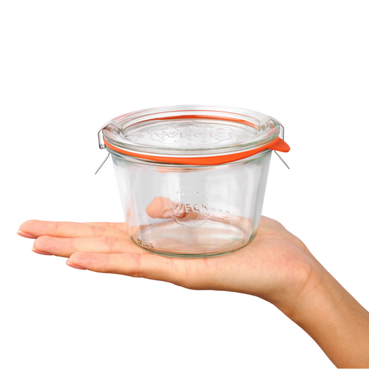 Прозрачная квадратная стеклянная банка Weck с оранжевым силиконовым уплотнителем и металлическими зажимами, удерживаемая женской рукой, подчеркивает надежность бренда для кухонного использования.