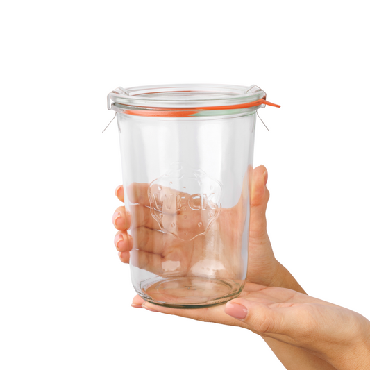Рука аккуратно держит прозрачную стеклянную банку WECK с оранжевым силиконовым уплотнением и стальными зажимами, подчеркивая высокое качество и уникальный дизайн с рельефным логотипом в форме клубники на лицевой стороне, идеальна для хранения продуктов.