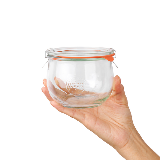 Ладонь поддерживает стеклянную банку WECK с оранжевой уплотнительной резинкой и металлическими зажимами, выпуклое гравированное лого, прозрачный фон.