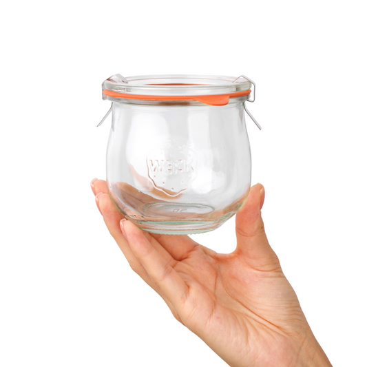 Ладонь аккуратно держит стеклянную банку WECK с оранжевой уплотнительной резинкой и стальными зажимами, виден рельефный логотип на прозрачной поверхности.
