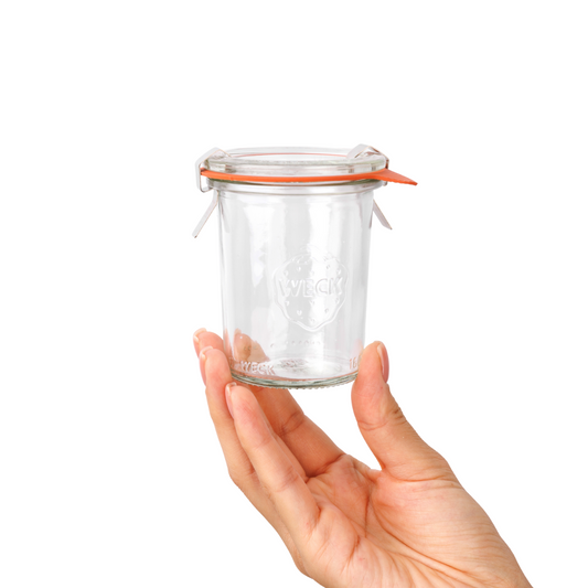 Рука держит прозрачную стеклянную баночку Weck с фирменной гравировкой и оранжевым силиконовым уплотнением, удобную для домашнего консервирования и хранения продуктов. Плотные стенки обеспечивают сохранность содержимого.