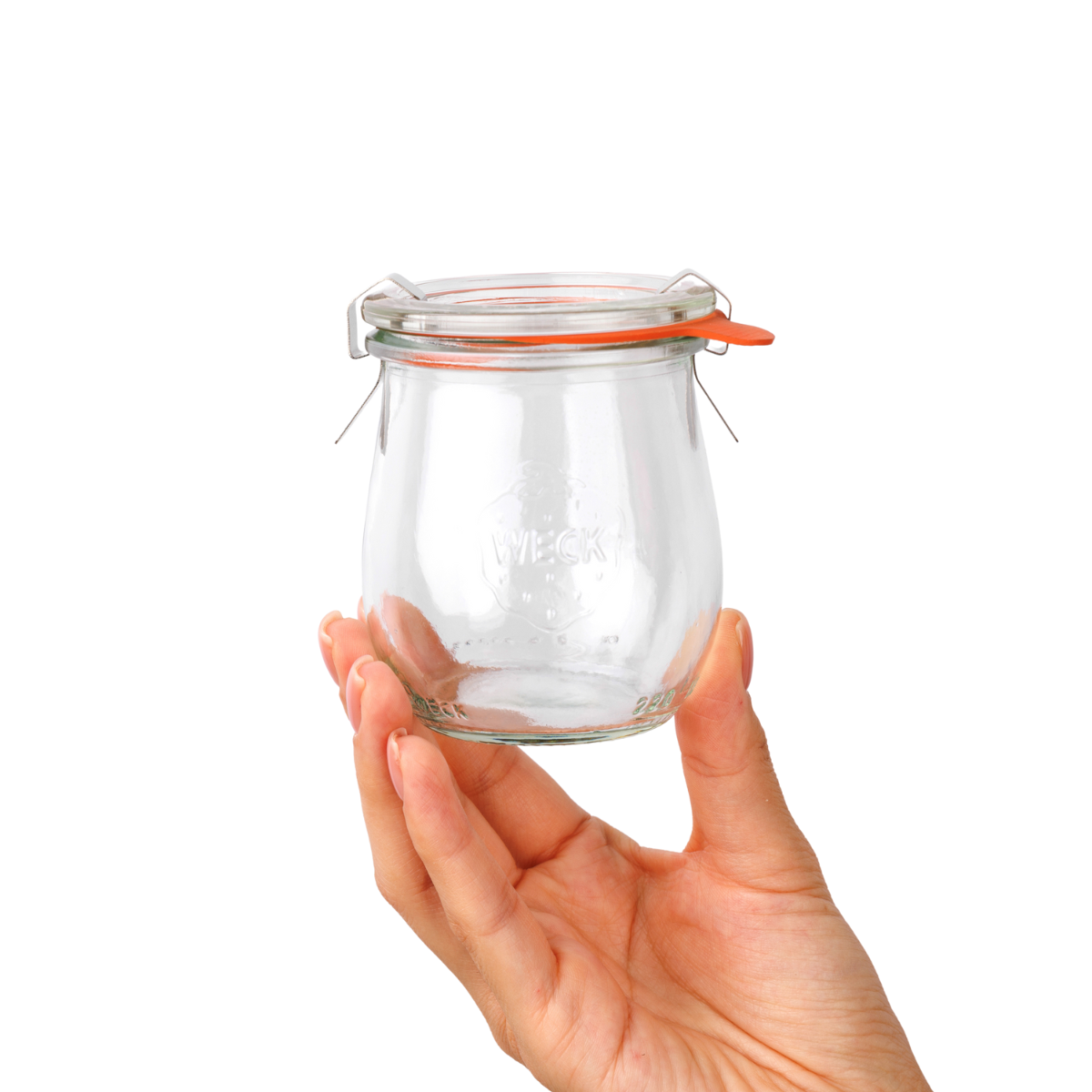 Рука нежно держит стеклянную банку WECK тюльпановой формы с прозрачными стенками, оранжевой уплотнительной резинкой и металлическими зажимами, с рельефным логотипом на передней стороне.