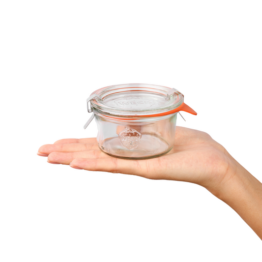 Рука держит круглую стеклянную банку Weck с оранжевыми зажимами и прозрачной крышкой, выгравированным логотипом на передней стороне, идеально подходящую для хранения домашних заготовок.