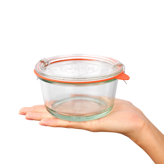 Стеклянная квадратная баночка Weck с крепкими стенками и прозрачной крышкой на руке, демонстрирующей ее компактный размер и удобную форму для хранения, с выгравированным логотипом, идеально подходящей для сервировки изысканных блюд.