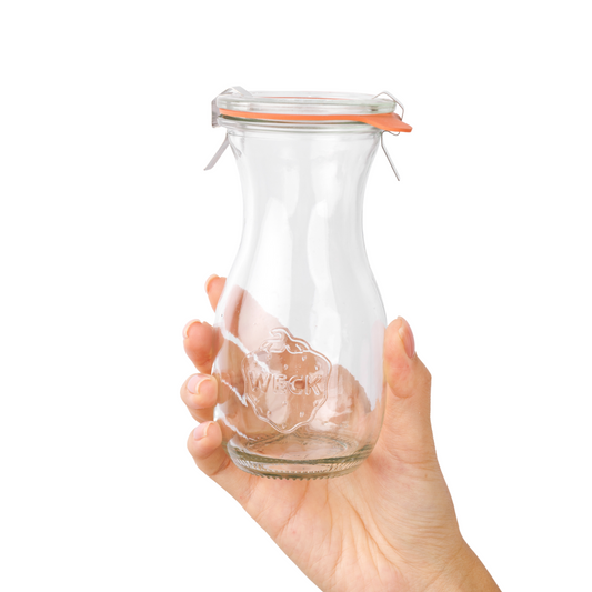 Рука нежно держит стеклянную банку WECK с узким горлышком, оранжевыми уплотнительной резинкой и зажимами, с гравировкой логотипа на фоне.