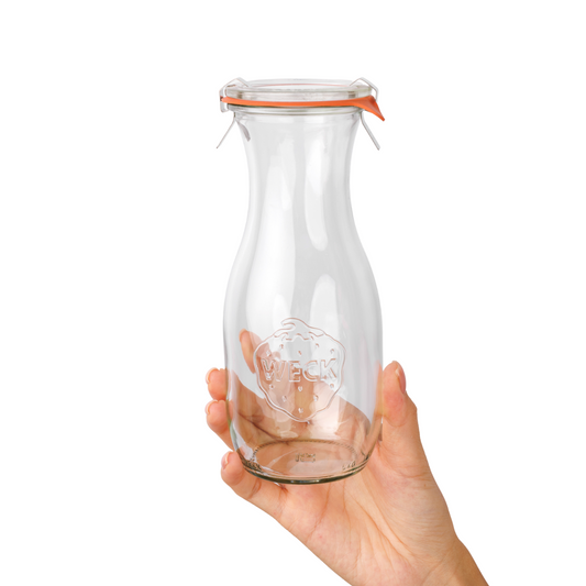 Рука держит высокую стеклянную бутылку WECK с оранжевой уплотнительной резинкой и металлическими зажимами, с гравировкой логотипа, на белом фоне.