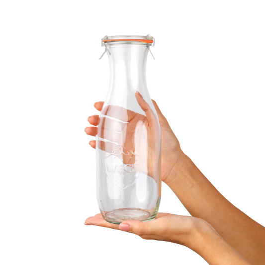 Рука держит стеклянную бутылку WECK с толстыми стенками и оранжевой резинкой, идеальную для хранения напитков и жидкостей, на белом фоне.