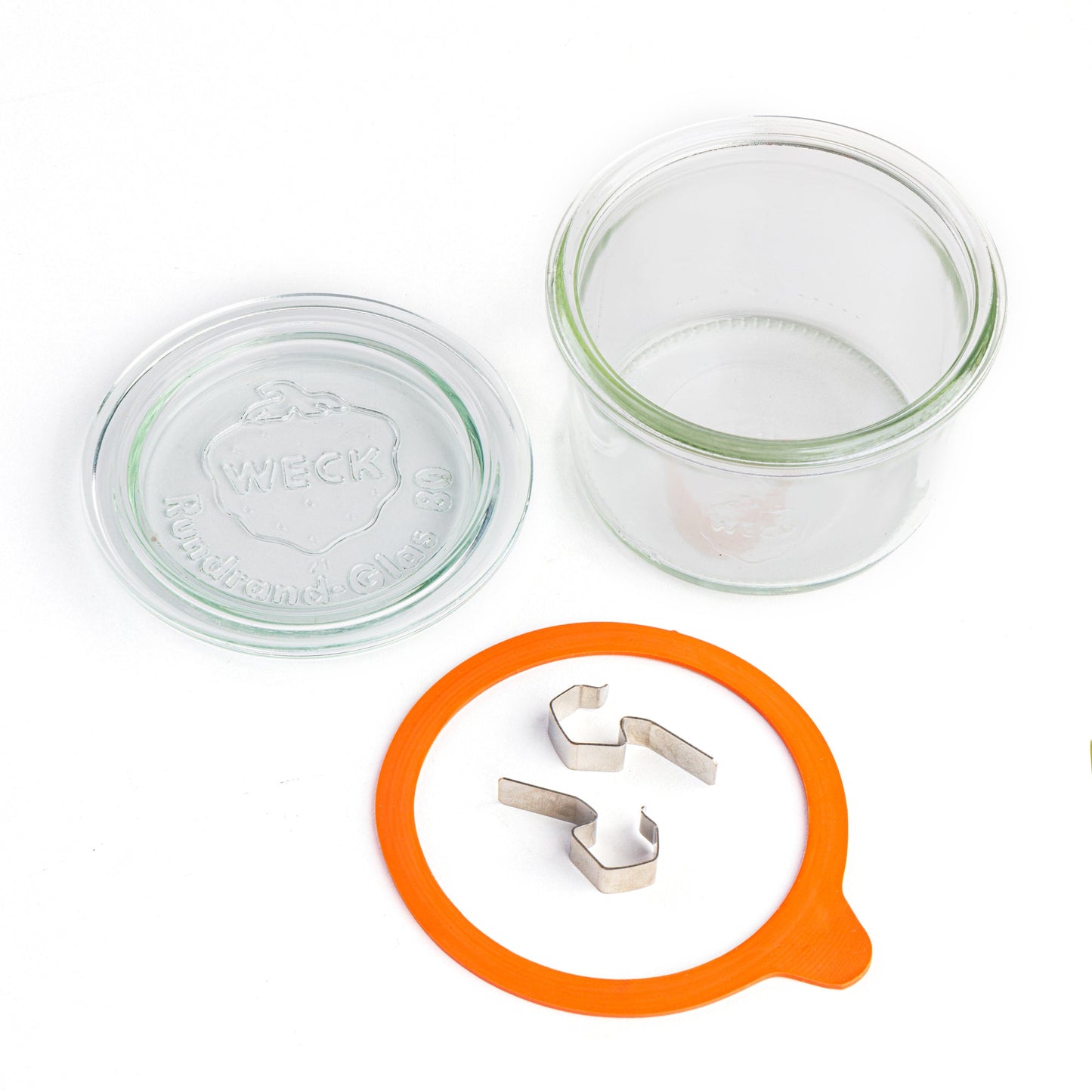 Компоненты стеклянной банки Weck: прозрачная крышка с эмблемой Weck, оранжевый силиконовый уплотнитель, стальные зажимы, изолированные на белом фоне.