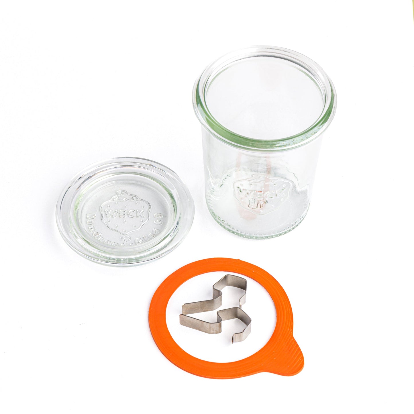 Круглая стеклянная банка Weck разобрана на компоненты: прозрачная крышка с выдавленным логотипом Weck, оранжевый силиконовый уплотнитель и два металлических зажима, на белом фоне.