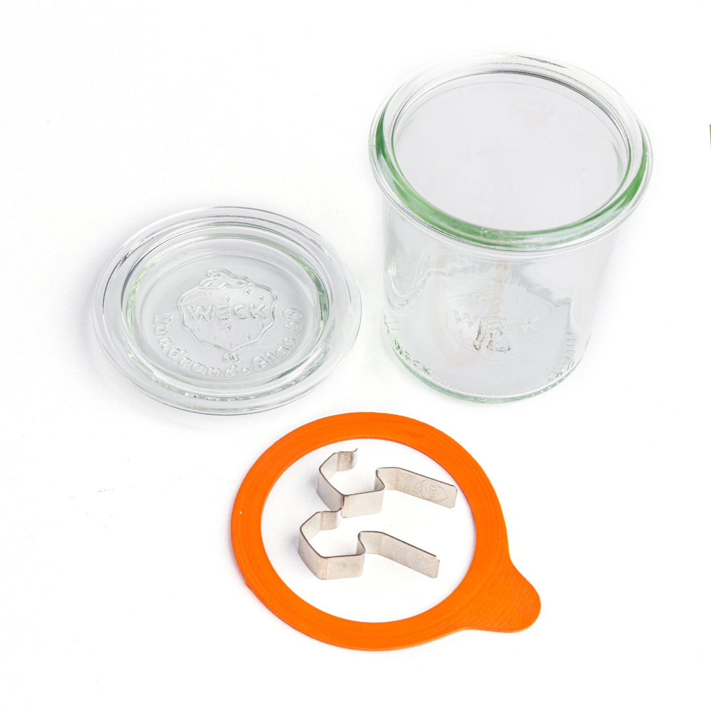 Компоненты круглой стеклянной банки Weck: прозрачная крышка с выдавленным логотипом, оранжевый силиконовый уплотнитель, и стальные зажимы на белом фоне.