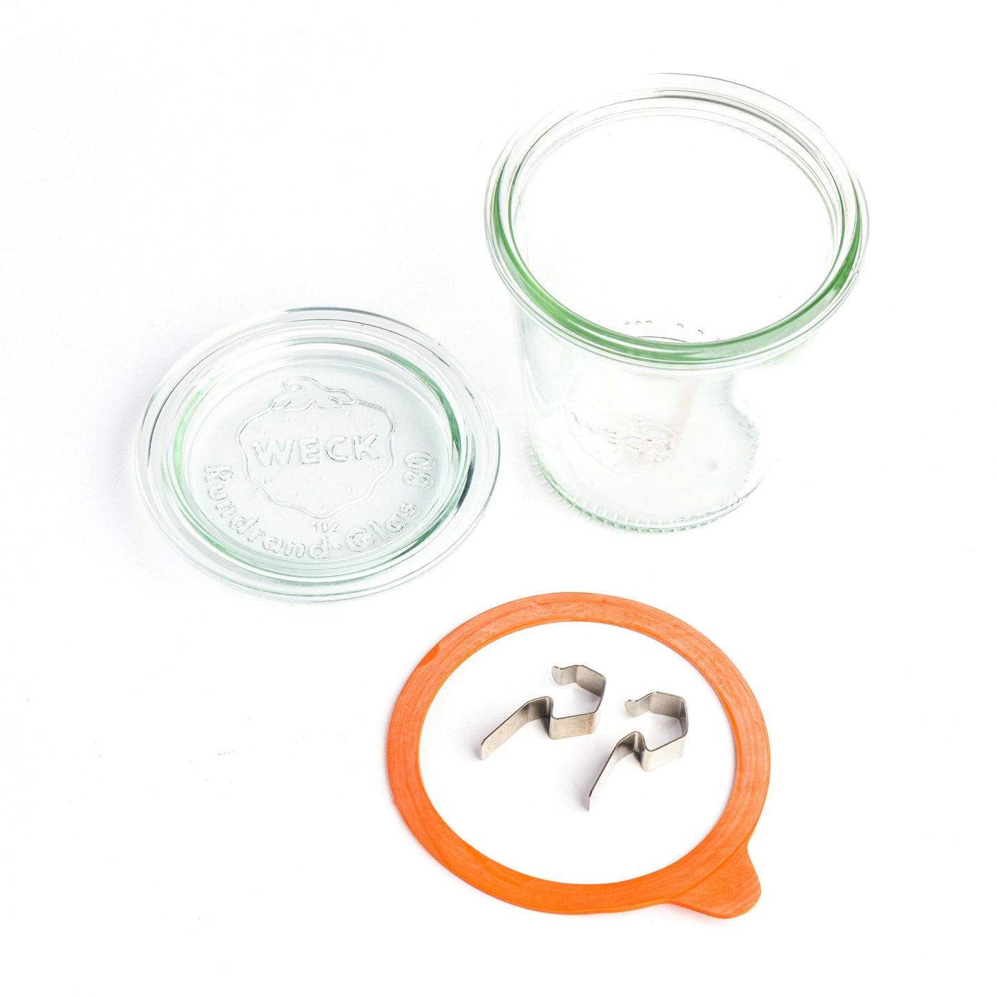 Круглая стеклянная банка Weck разобрана на компоненты: прозрачная крышка с гравировкой логотипа, оранжевый силиконовый уплотнитель и зажимы, на белом фоне.