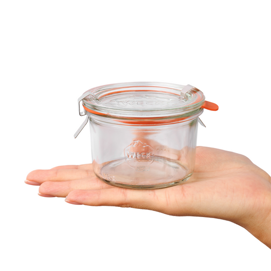 Прозрачная стеклянная банка Weck с плотно закрывающейся оранжевой крышкой и металлическими зажимами, удерживаемая женской рукой, подходит для элегантного сервирования и хранения продуктов.