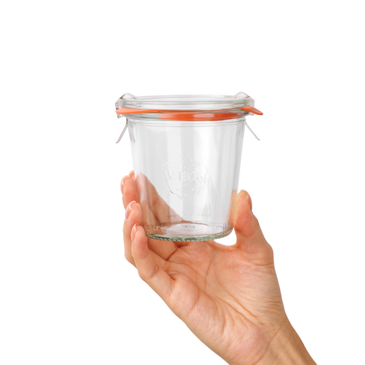 Квадратная стеклянная банка Weck с оранжевой уплотнительной резинкой и стальными защелками, демонстрируемая в женской руке, подчеркивает качество и универсальность использования для хранения продуктов.